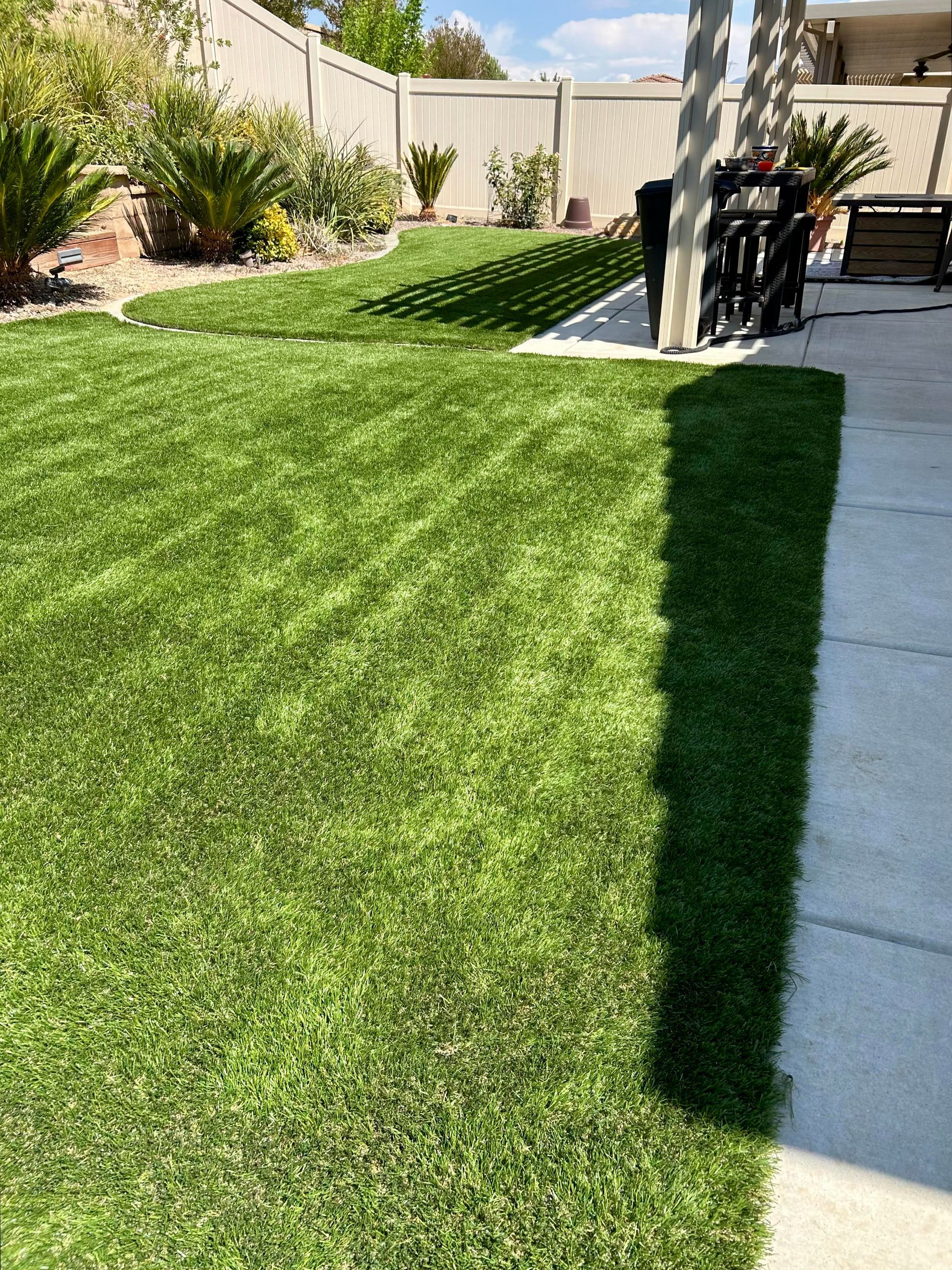 Sunny backyard with green artificial turf, white fence, and concrete patio in the foreground