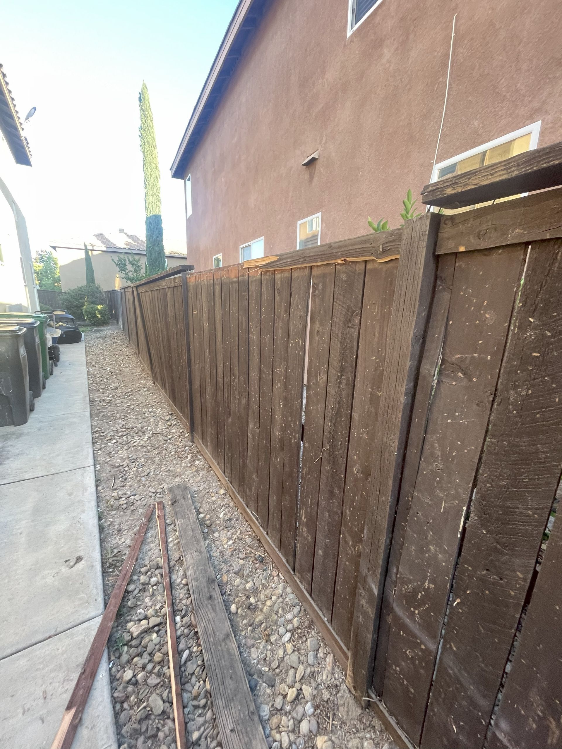 Brown wooden fence along a gravel path beside a light brown stucco building.