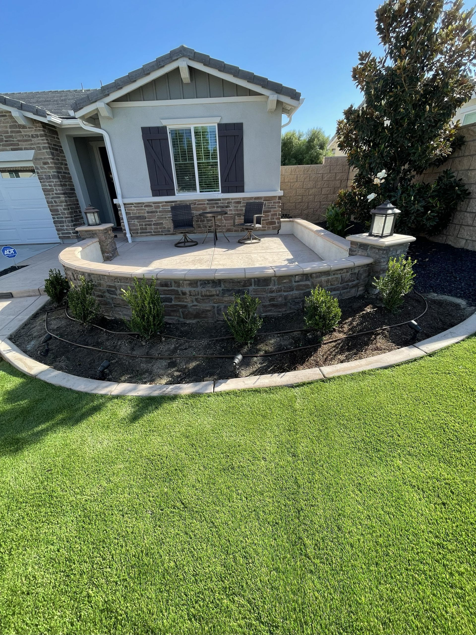 Front yard with a curved stone retaining wall, concrete patio, and a house exterior under a clear blue sky