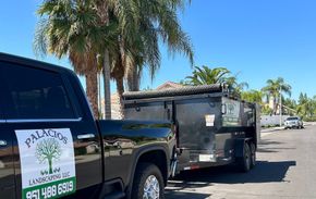 A black truck with a tree on the back is parked next to a dumpster.