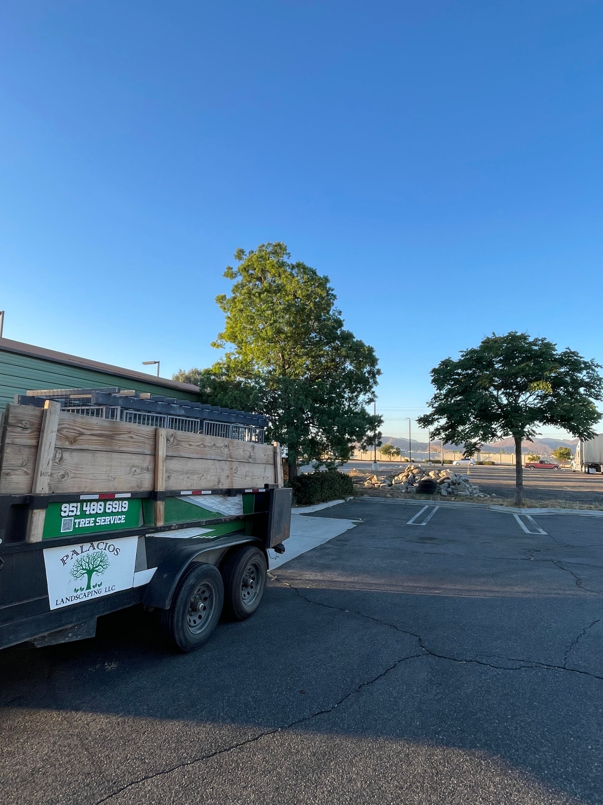 Trailer loaded with building materials parked in a parking lot near trees under a clear blue sky. The trailer has green and white logos.