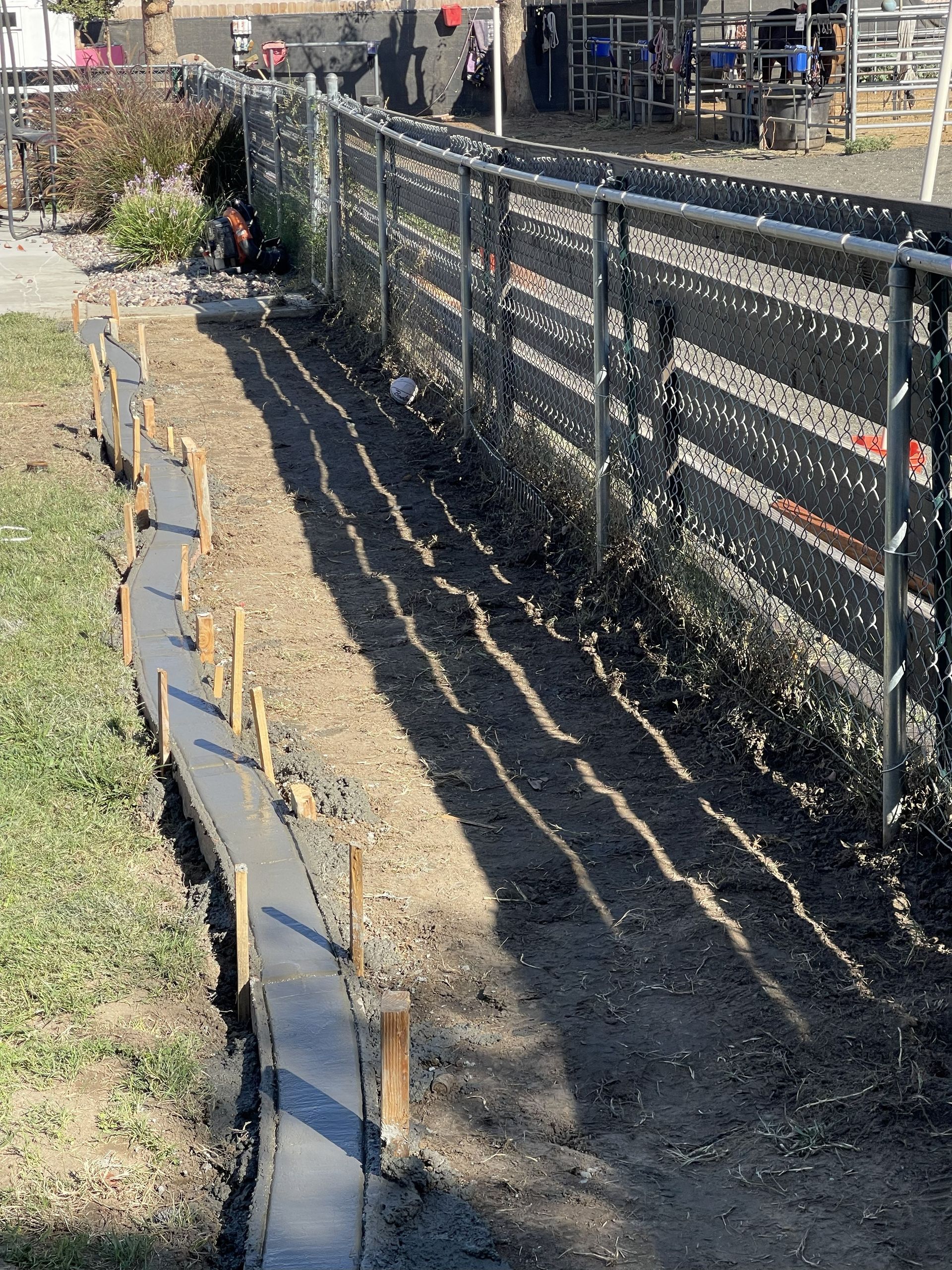 A concrete curb under construction next to a chain-link fence, with wooden stakes and shadows.