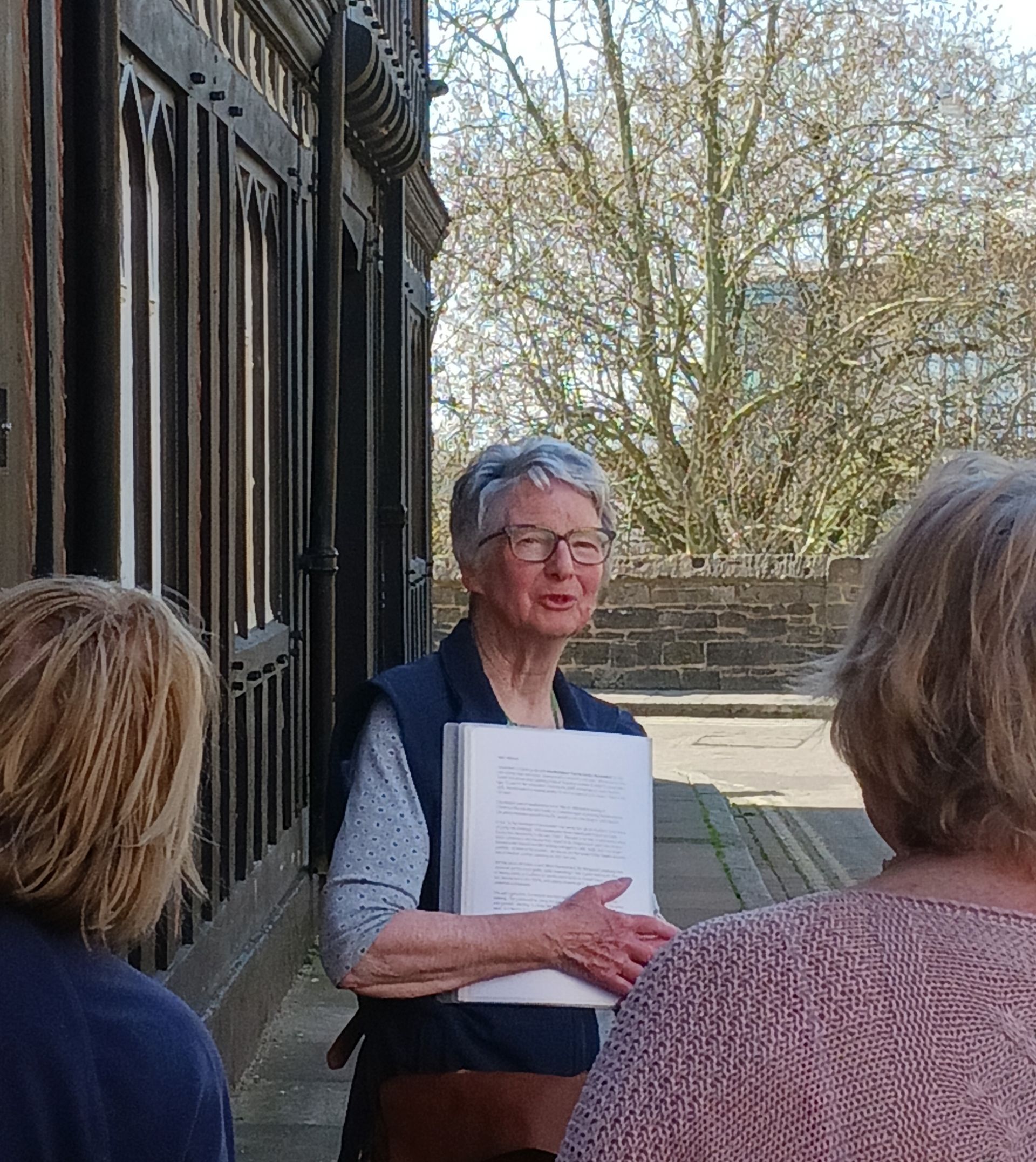 A woman is standing in front of a building holding a book and talking to a group of people.