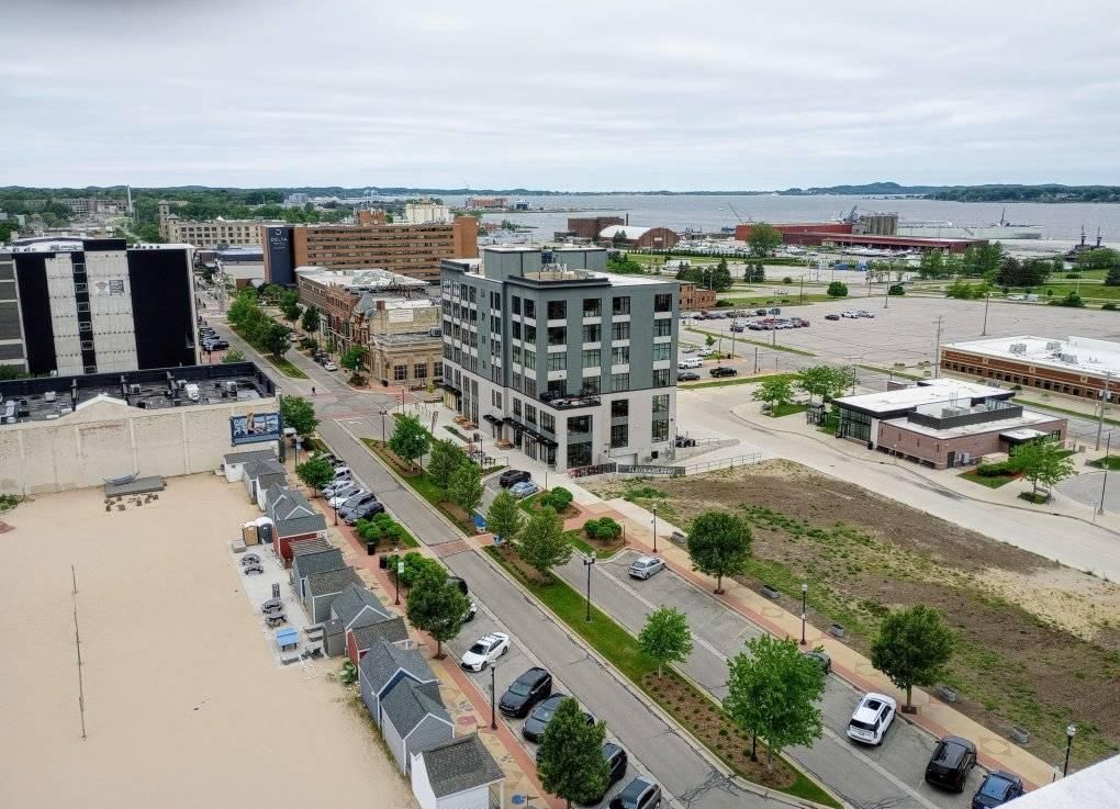 Aerial view of a waterfront city block with roads, modern buildings, parking lots, and a bay in the distance