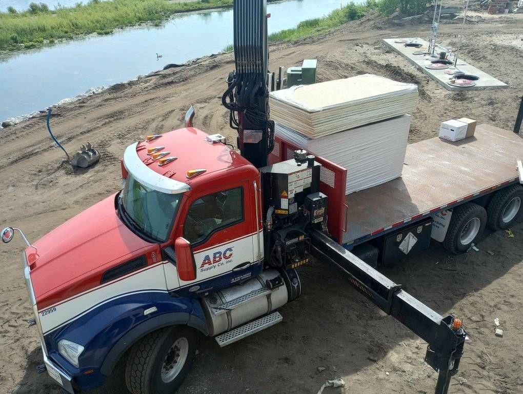 Red flatbed truck with crane hauling stacked white materials beside a canal.
