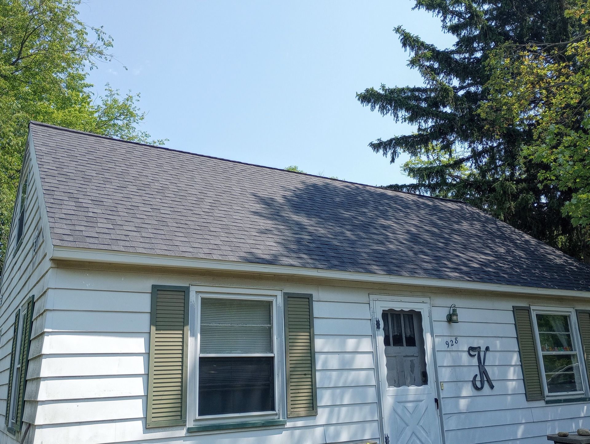 Small white house with gray shingled roof, green shutters, and a front door under trees.