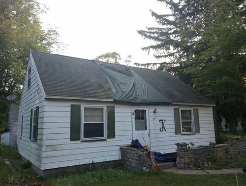 Small white house with damaged dark roof, front porch, and trees around it