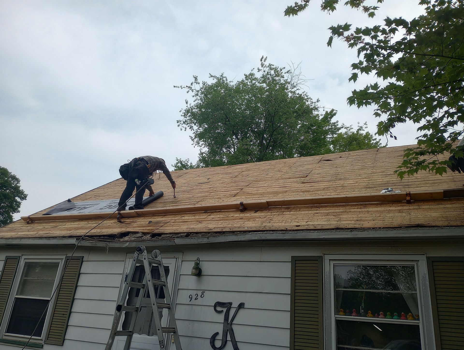 Worker replacing roof shingles on a house while standing near a ladder under a cloudy sky