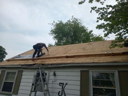 Worker on a house roof replacing shingles, with a ladder leaning against the siding.