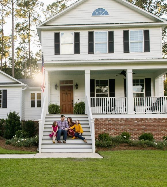 Three people sit on the front steps of a white house with a porch and brick foundation.