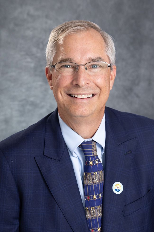 Man with glasses smiles, wearing a blue striped shirt and patterned tie, outside.
