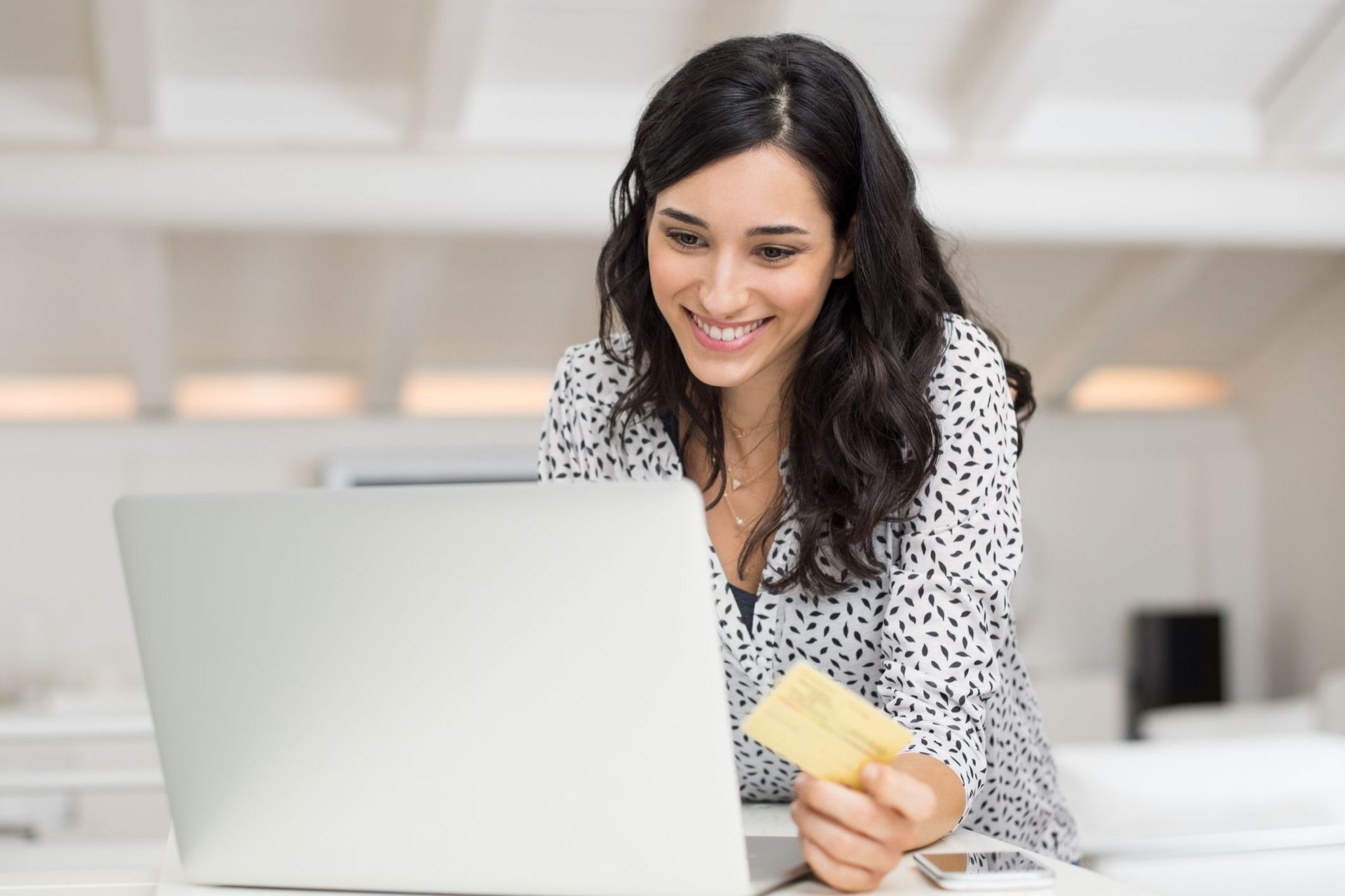 Woman with dark hair smiles, looking at a laptop while holding a gold credit card.