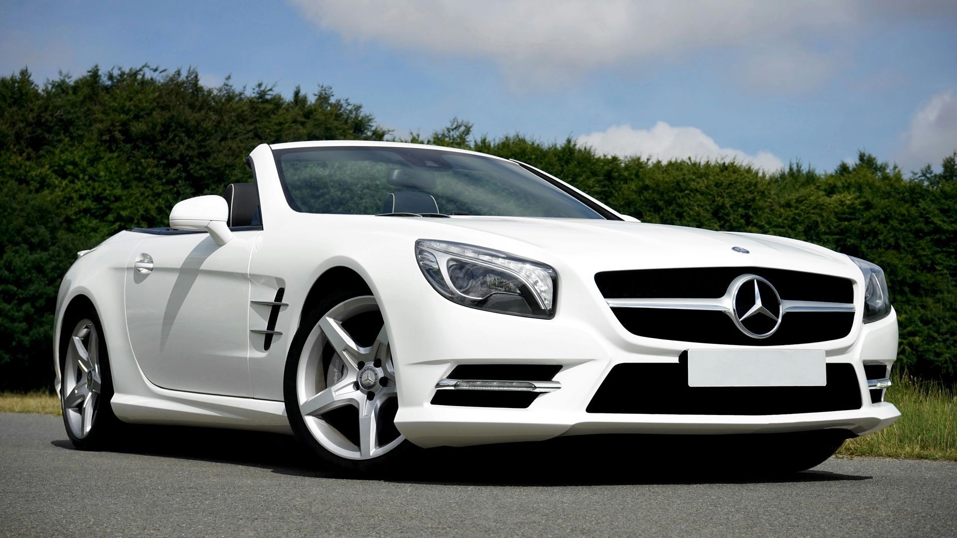 White Mercedes convertible on a paved road, with trees in the background, against a blue sky.