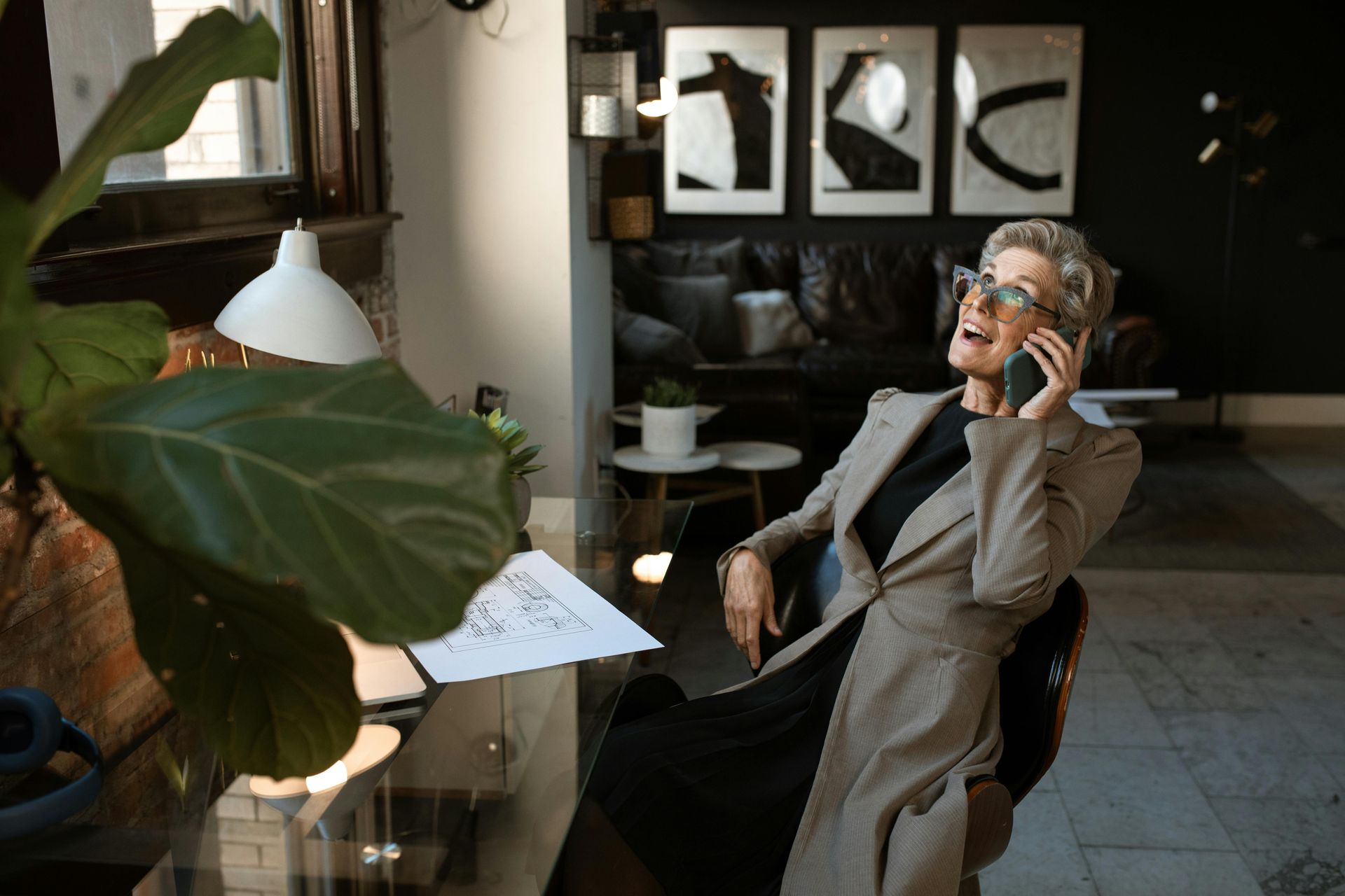 Woman in blazer, talking on phone, laughing, sitting at desk in stylish room.