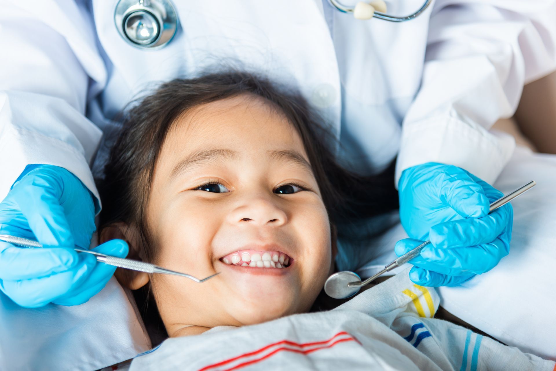 A smiling boy with healthy teeth holds a plush tooth and a toothbrush on a blue isolated background. Dentist using tools in a child's mouth while the child smiles in a dental chair
