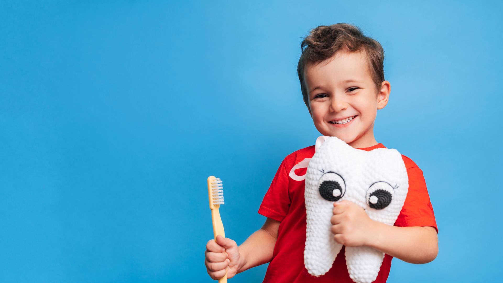 A smiling boy with healthy teeth holds a plush tooth and a toothbrush on a blue isolated background.