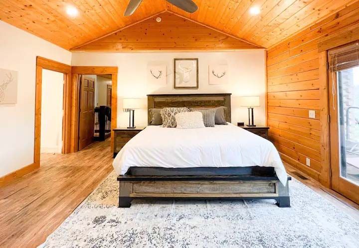 Bedroom with wooden walls, vaulted ceiling, bed with gray headboard, rug, doorway.