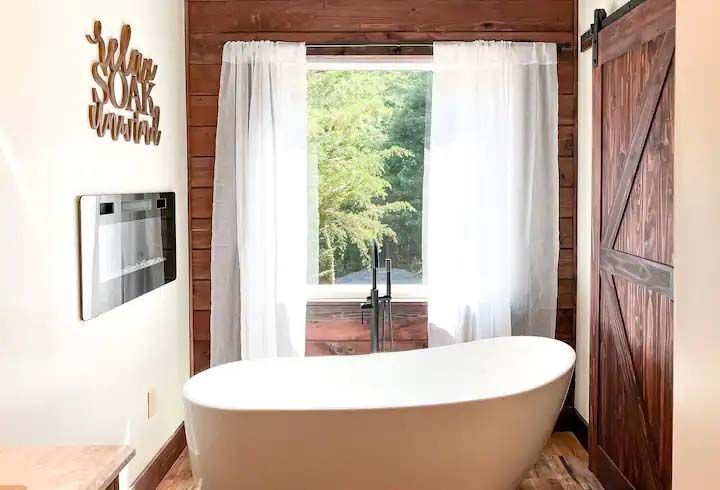 Bathroom with a white soaking tub, window with sheer curtains, and a wooden sliding door.