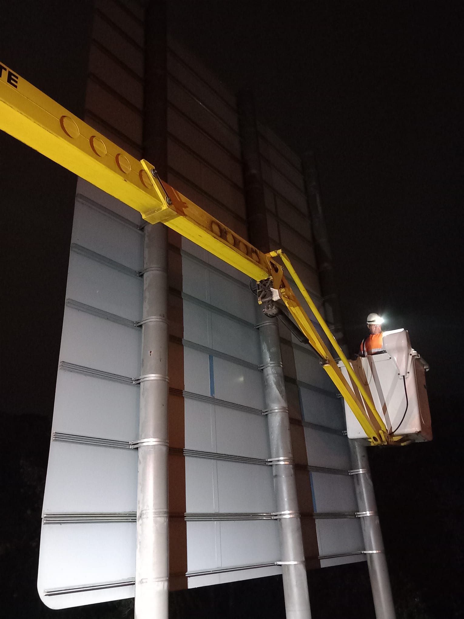 Yellow boom lift raising a worker basket beside a tall gray metal wall at night