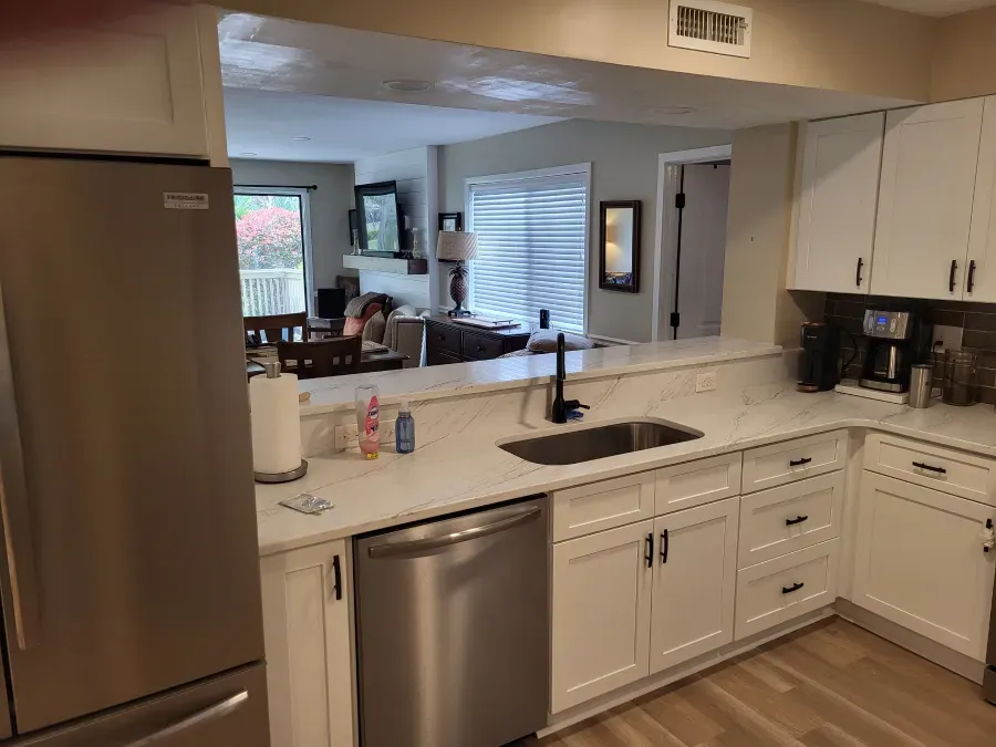 Modern kitchen with white cabinets, stainless steel appliances, and a view into a living room. A countertop sink and pass-through window are visible.