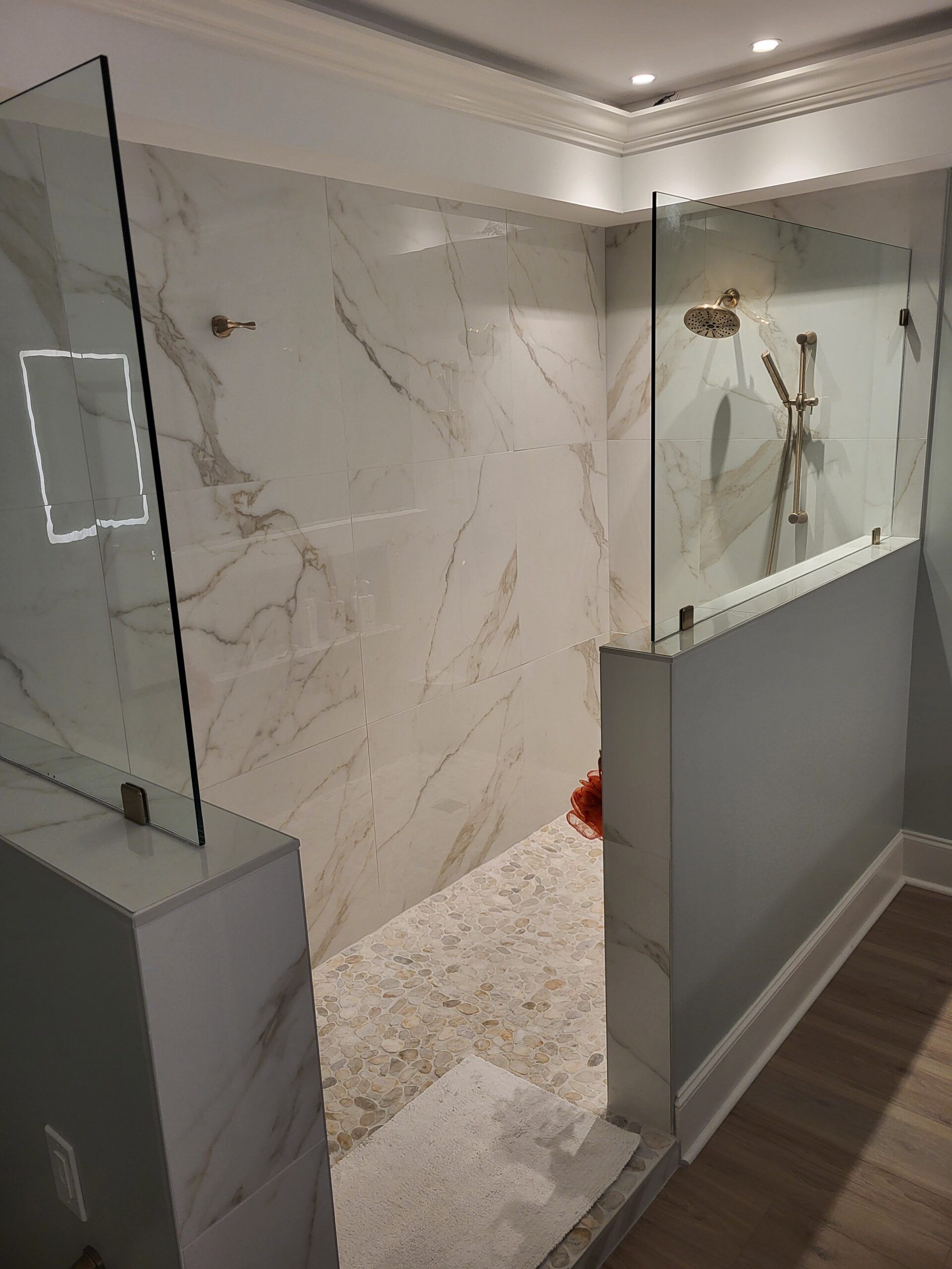 A modern shower with marble-like walls, glass panels, and gold shower fixtures. The entrance is partially blocked by a wall.