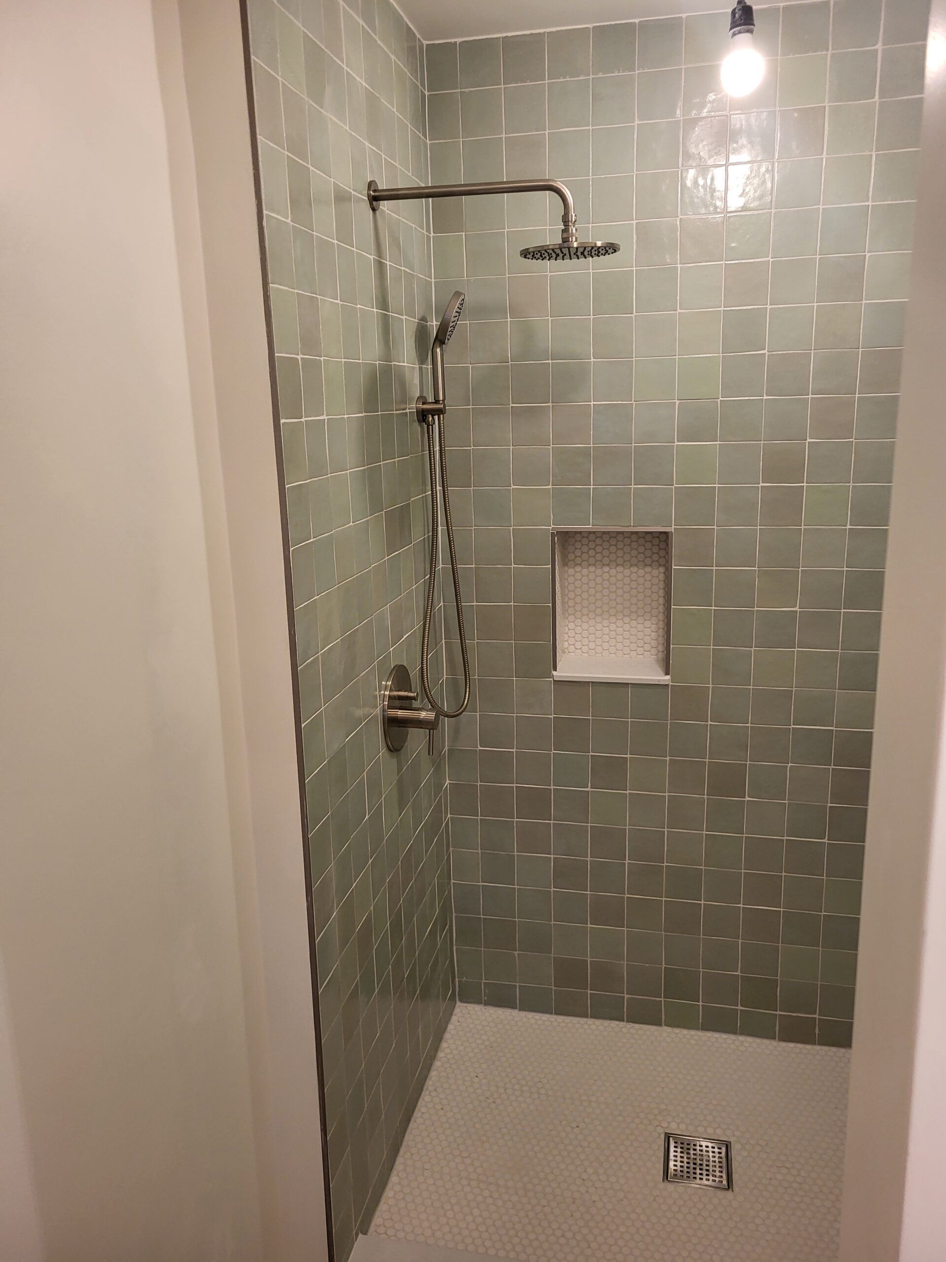 Green tiled shower with chrome fixtures and a built-in shelf. White floor with a drain.