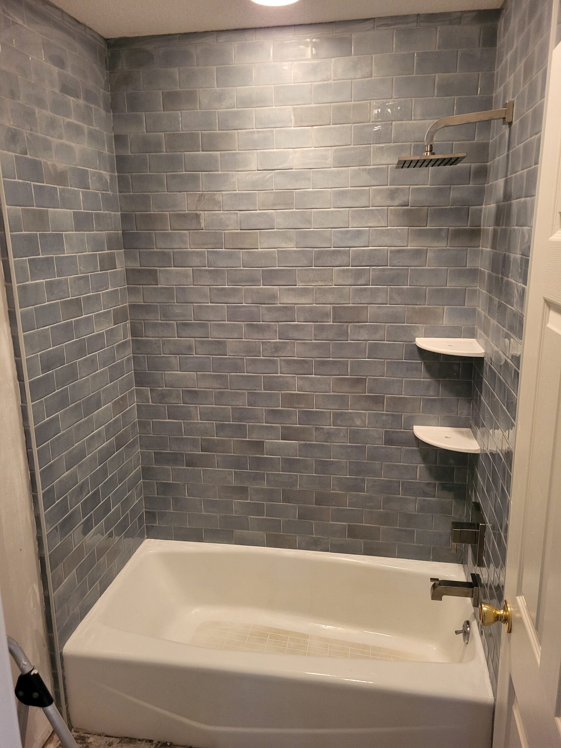 Blue tiled shower with white tub, two rounded shelves, and gold showerhead.