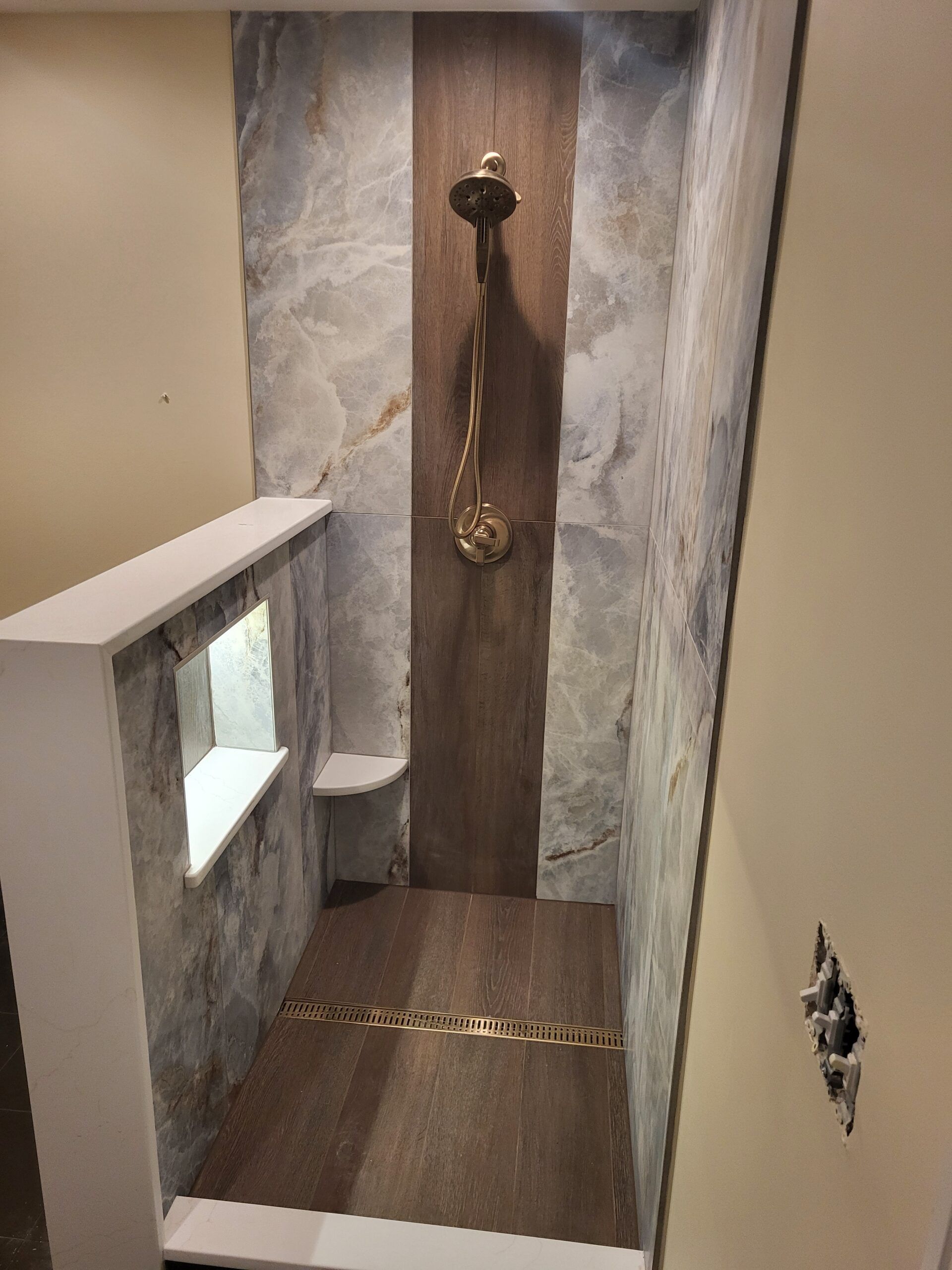 Shower with brown and gray stone tile walls, a dark wood accent, and gold fixtures. A small window with a shelf is visible.