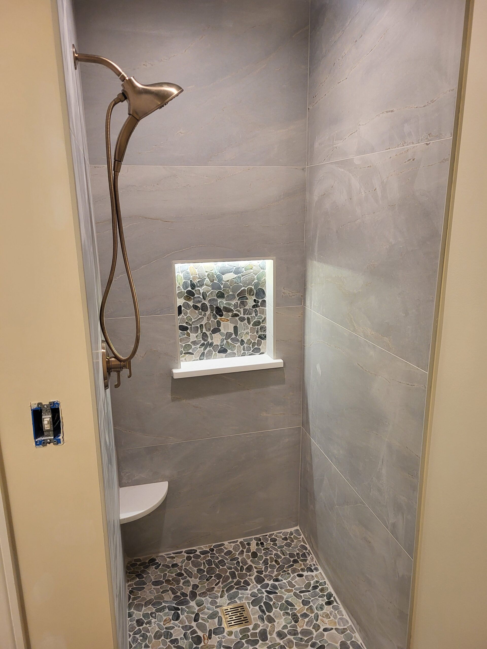 Small shower with gray walls and pebble floor, featuring a bronze showerhead and a built-in shelf.