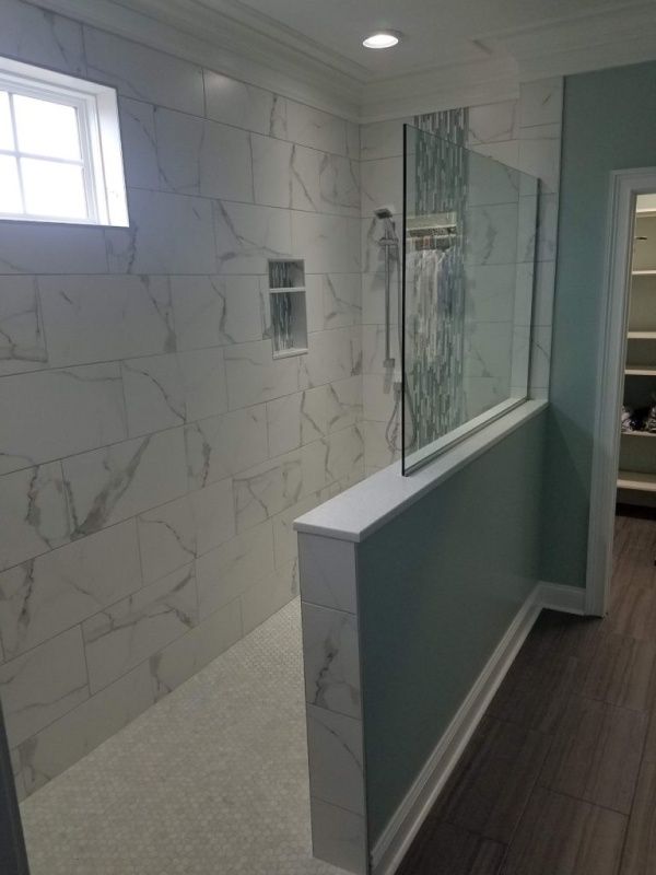 A modern bathroom with white marble-look tiles and a glass shower door. A half-wall is painted teal, and there's a small window.