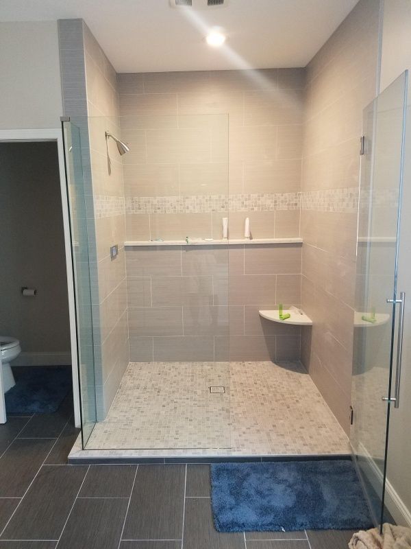 Modern bathroom with a walk-in shower featuring gray tiles, glass doors, and a built-in bench. A blue rug sits on the floor.