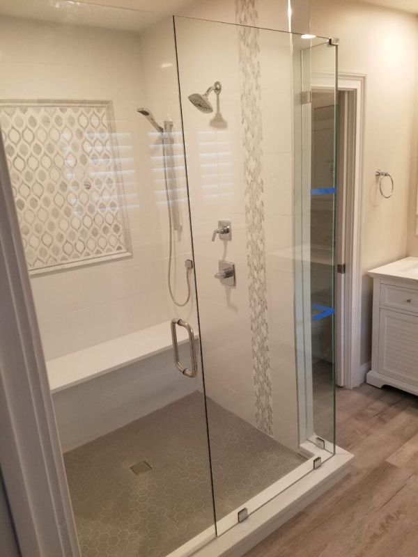 A modern bathroom with a glass shower, white walls, and light wood-look flooring. The shower features a bench, multiple showerheads, and decorative tile.