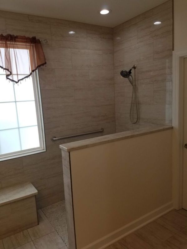 Accessible bathroom with beige tiled shower and a clear glass partition. Features a window with a bench seat, a grab bar, and a showerhead.