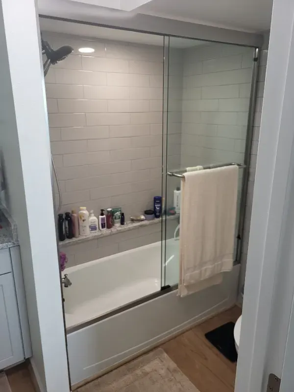 A modern bathroom with a white bathtub, glass shower doors, and light gray tiled walls. A towel hangs on the shower door.