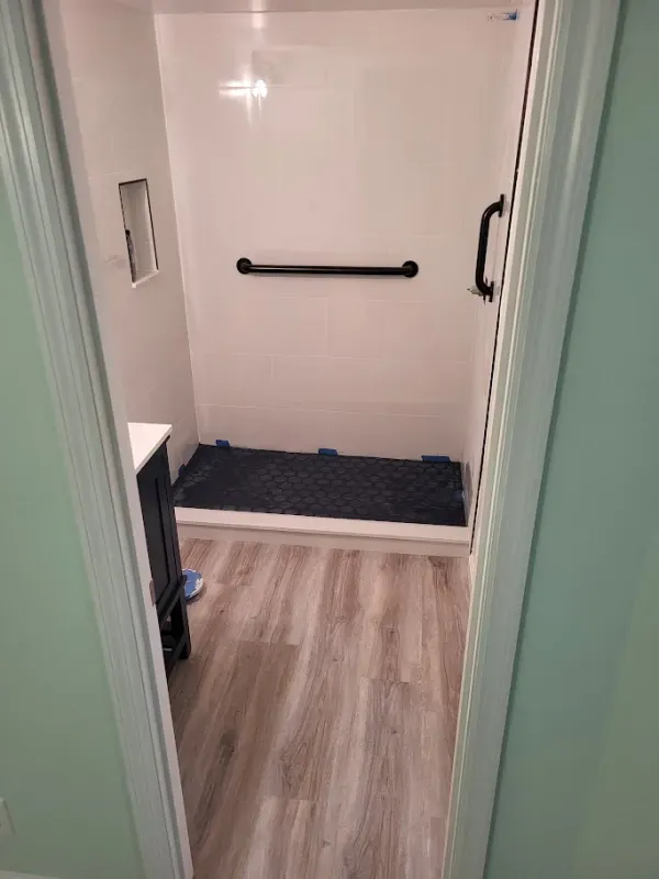 Bathroom interior featuring a white walk-in shower with a black grab bar and a non-slip mat. Light wood-look flooring.