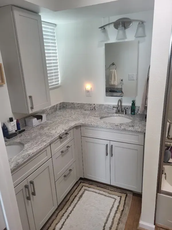 White bathroom with a granite countertop, two sinks, white cabinets, and a light fixture over a mirror. A window with blinds is visible.