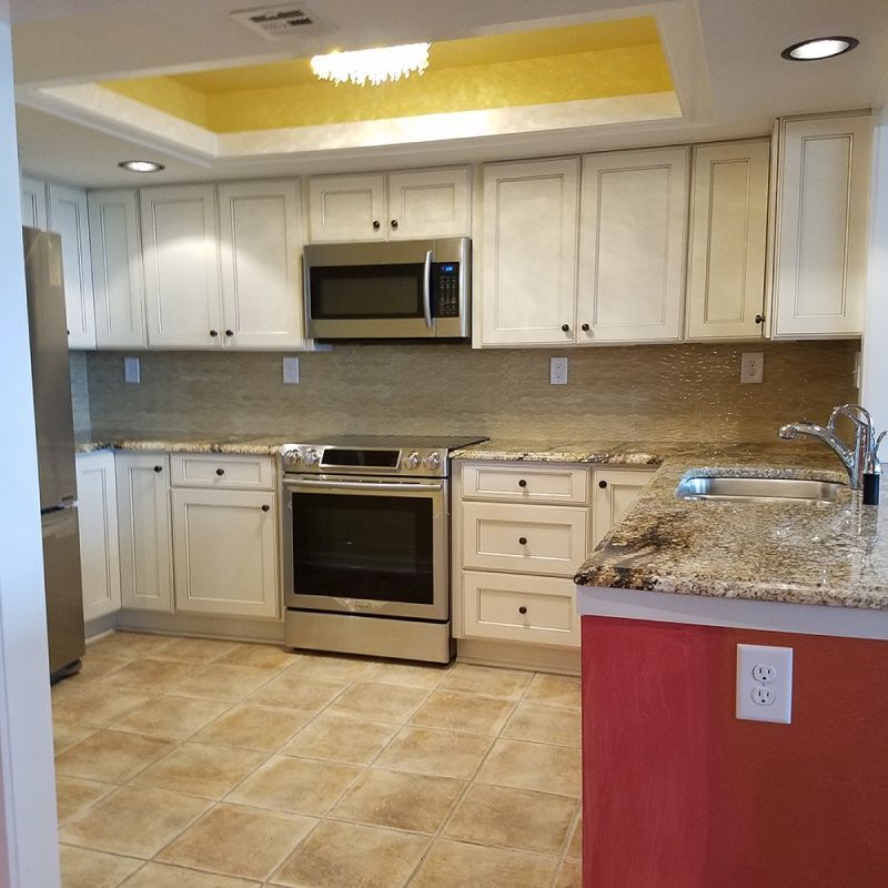 A renovated kitchen with white cabinets, stainless steel appliances, granite countertops, and tan tile floors. Red cabinet end accent.
