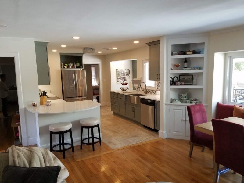 Open-concept kitchen with white countertops, gray cabinets, and wooden floors. A breakfast bar with stools is on the left, a dining table on the right.