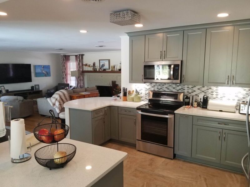 A modern kitchen with light blue cabinets, white countertops, and stainless steel appliances opens to a living room.
