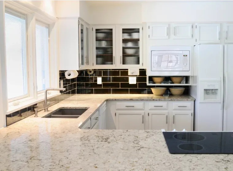 A bright, white kitchen with a black tile backsplash. A stainless steel sink, microwave, and refrigerator are present. The countertops are light-colored with a cooktop visible in the foreground.