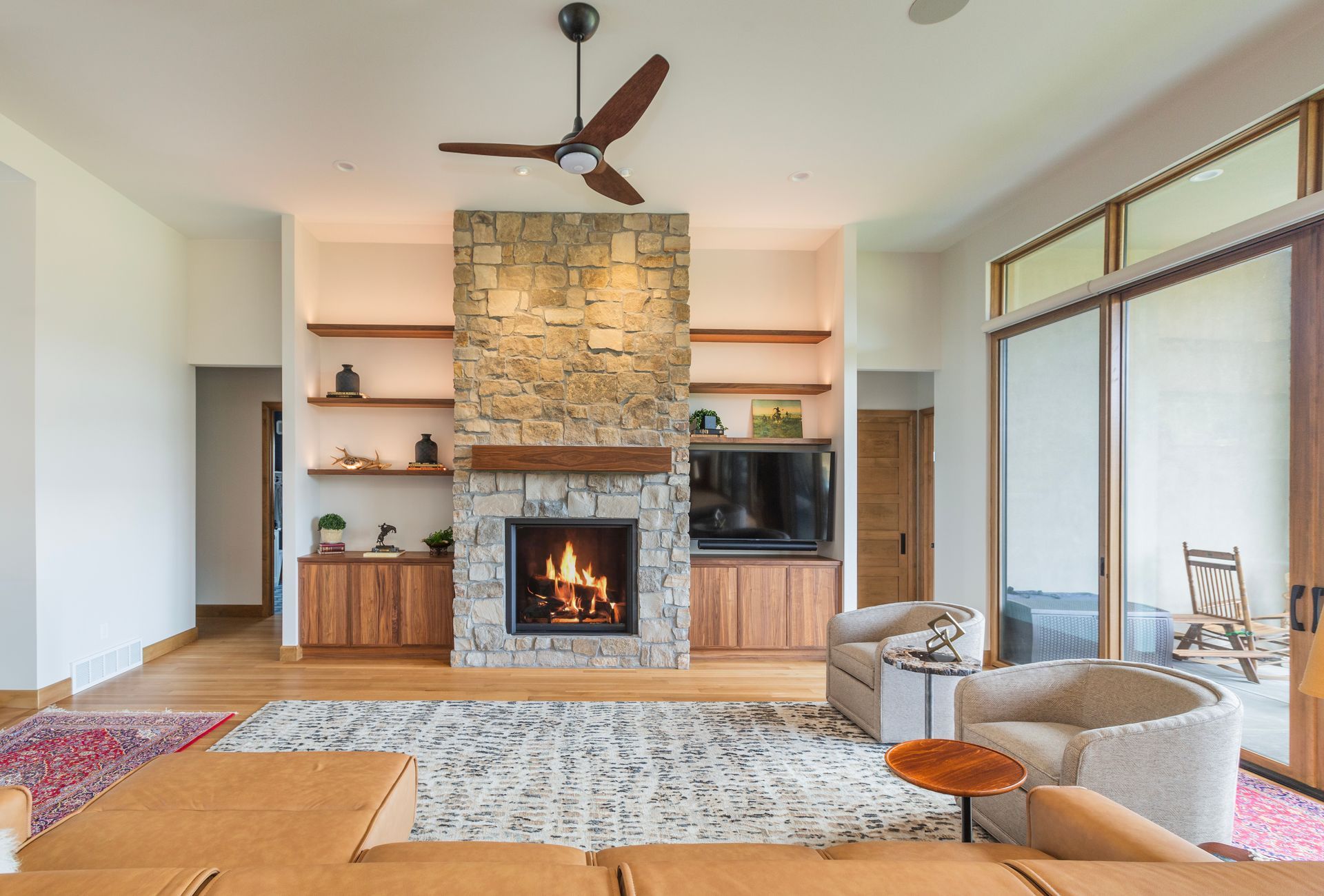 Modern living room with stone fireplace, built-in shelves, large windows, and ceiling fan