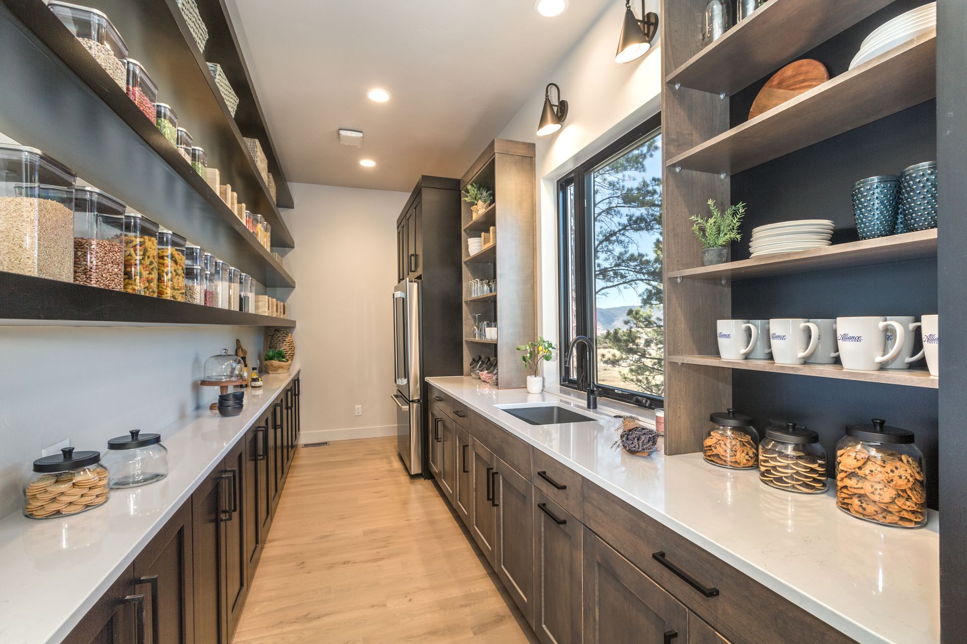 Modern pantry with open shelving, wood floor, white counters, and jars of dry goods lining both sides.