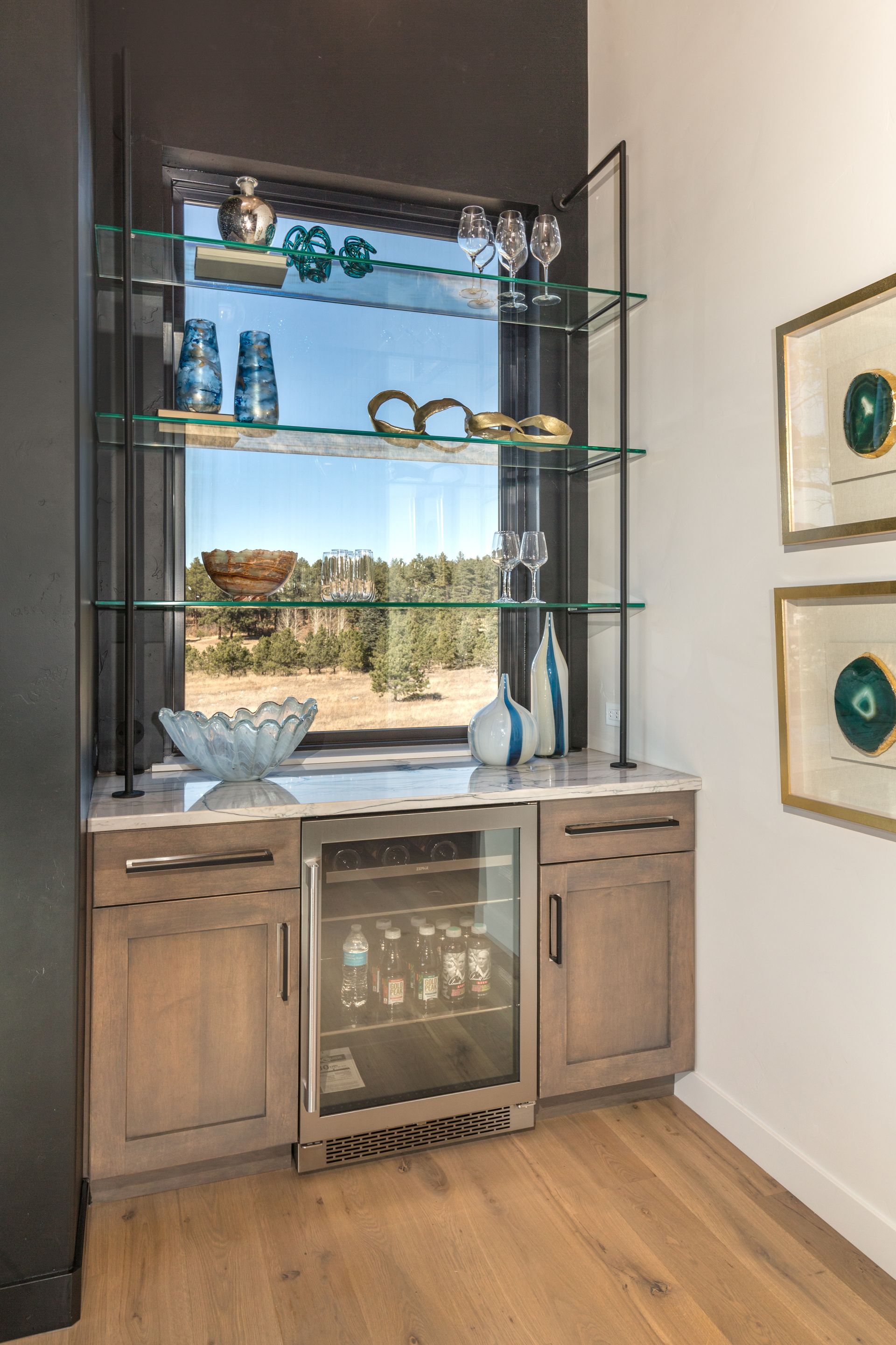 Built-in wooden cabinet with glass shelves, decor, and wine cooler beside a bright window.