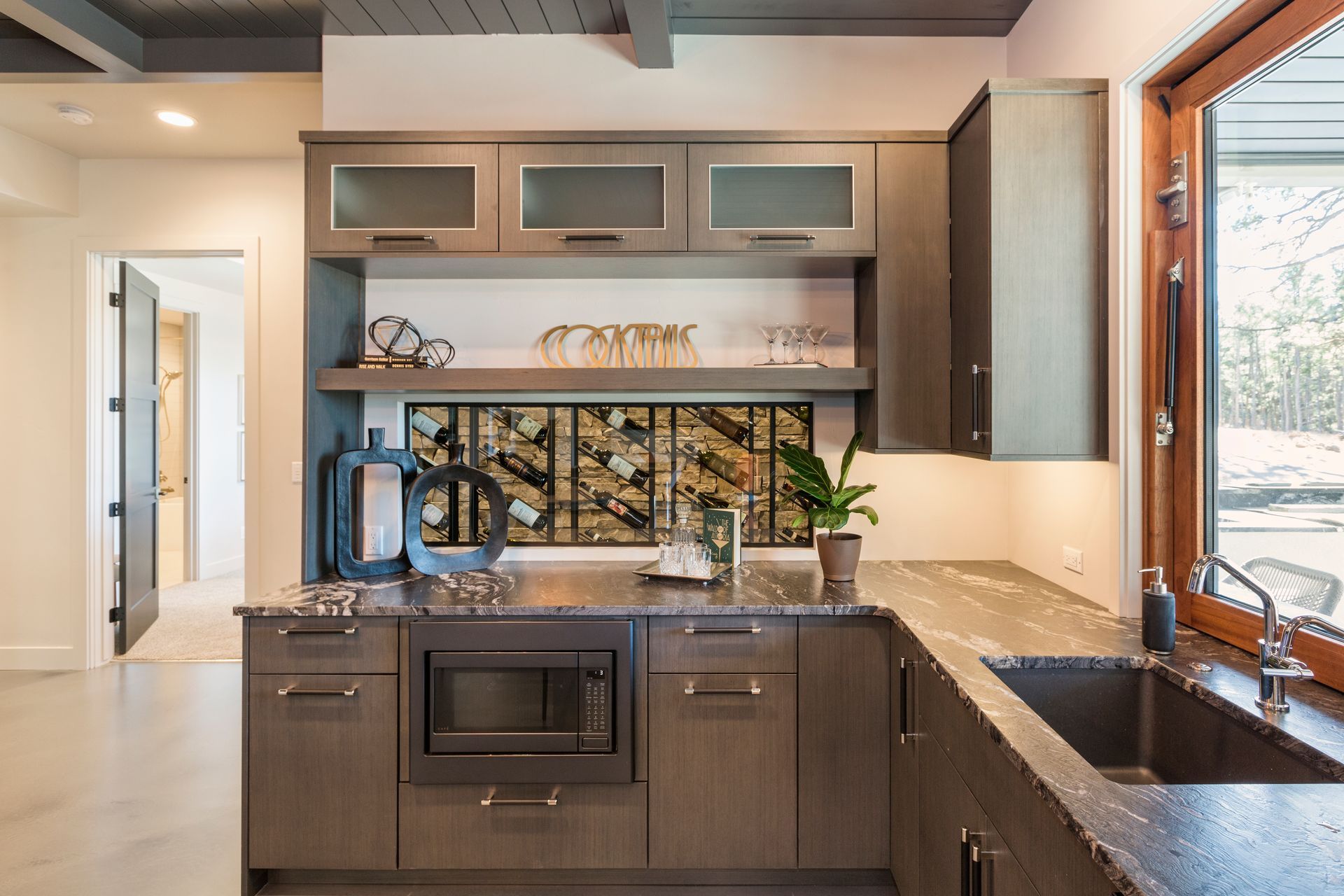 Modern kitchen with gray cabinets, black sink, built-in oven, and large window letting in daylight