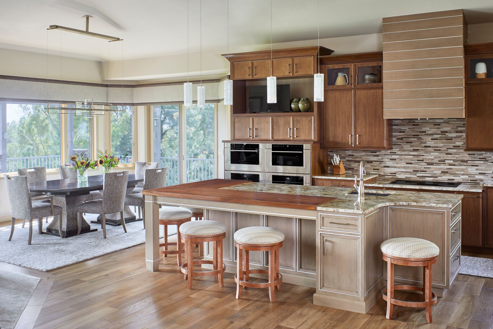 Open-plan kitchen and dining area with wood cabinets, island stools, and large windows overlooking greenery