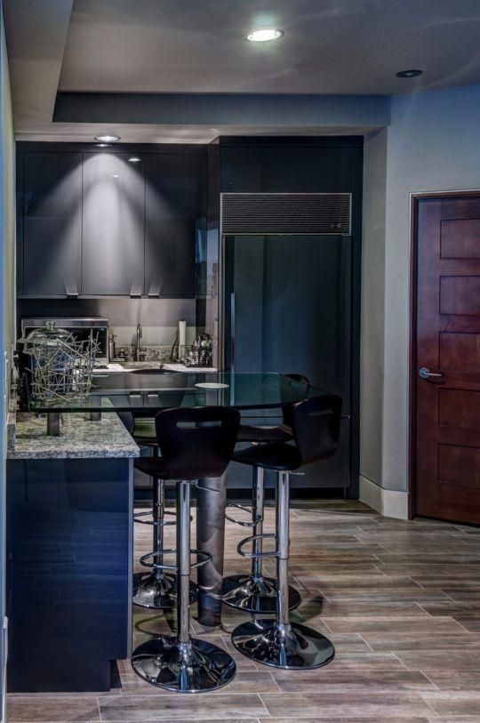 Modern kitchen with granite island, two bar stools, stainless fridge, and wooden door under recessed lighting