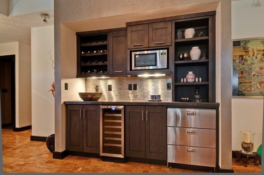 Built-in home bar with wood cabinets, wine cooler, microwave, and decorative shelves in a tiled alcove