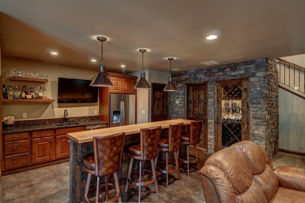 Warm home bar with wood cabinets, stone fireplace, island stools, and beige leather couch in an open basement.