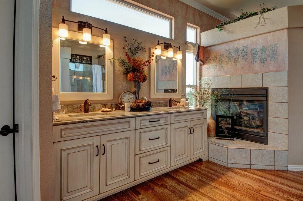 Bathroom vanity with double sinks, mirrors, wall sconces, and a tiled fireplace beside hardwood floors