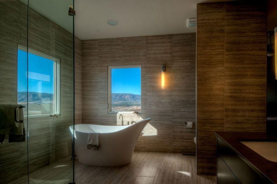 Modern bathroom with freestanding tub, large window, glass shower, and wood-paneled walls overlooking mountains
